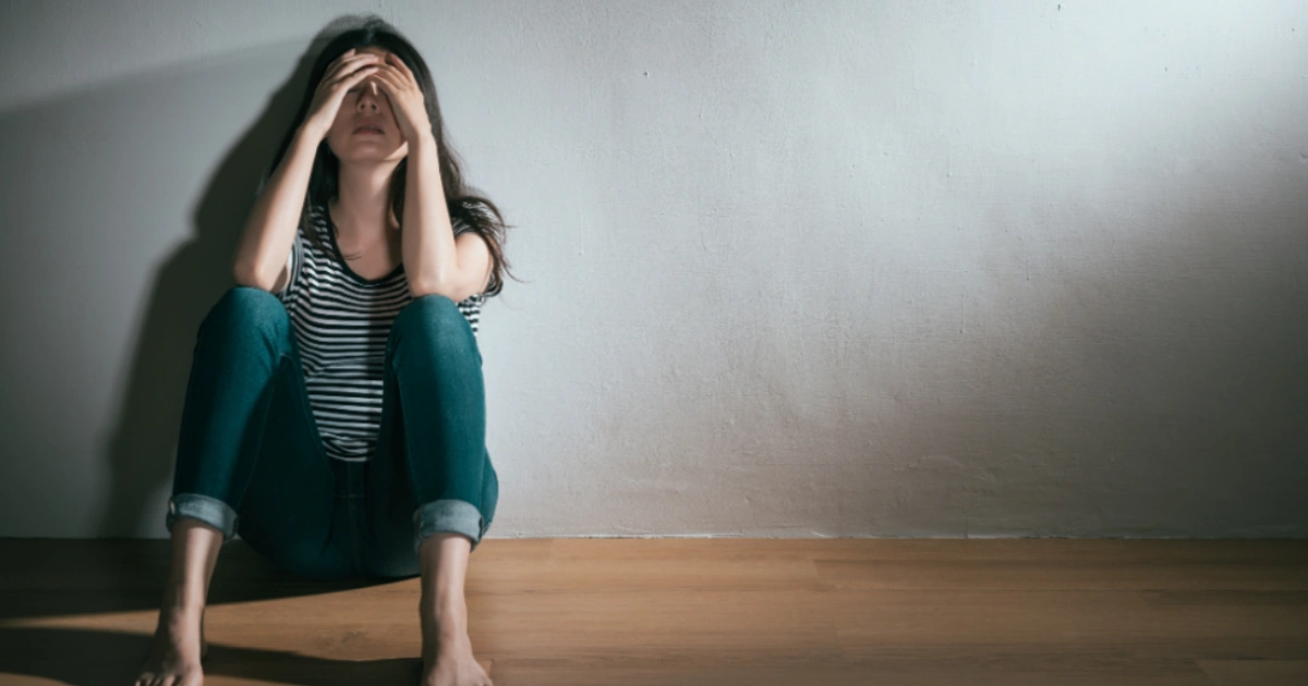 Woman sitting on the floor against a wall with her hands over her face in distress