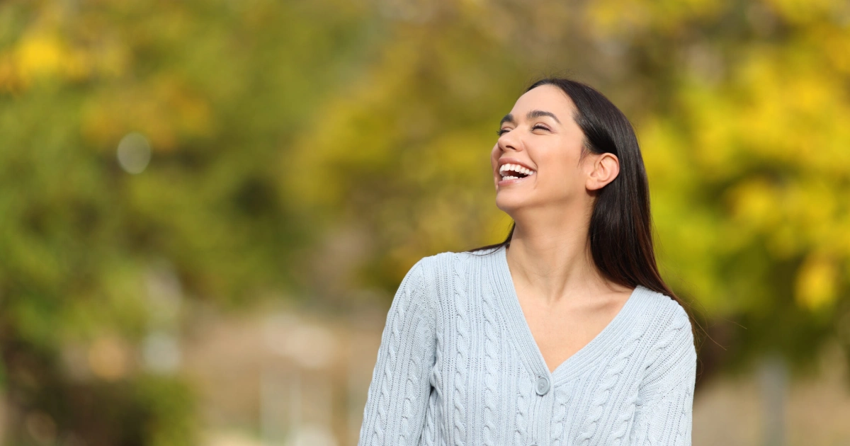 Smiling woman outdoors wearing a light sweater with autumn trees in the background