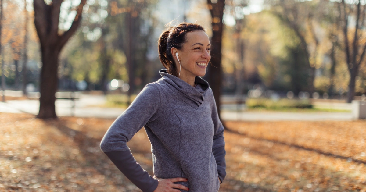 A woman in a gray long-sleeve top stands outdoors with hands on hips, smiling and wearing earphones. Fall leaves cover the ground and trees with autumn foliage are in the background.