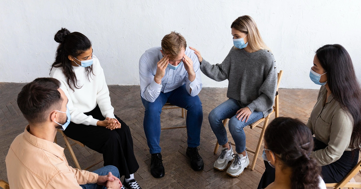 A group of six people wearing masks sits in a circle. One man in the center appears distressed, holding his head, while a woman beside him comforts him by touching his shoulder. The others watch supportively.