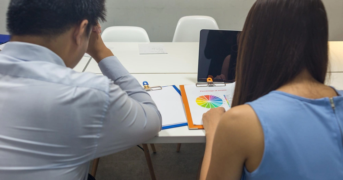 Two people sit at a table reviewing documents and colorful charts on clipboards; a tablet and notepad are also on the table, with both individuals viewed from behind.