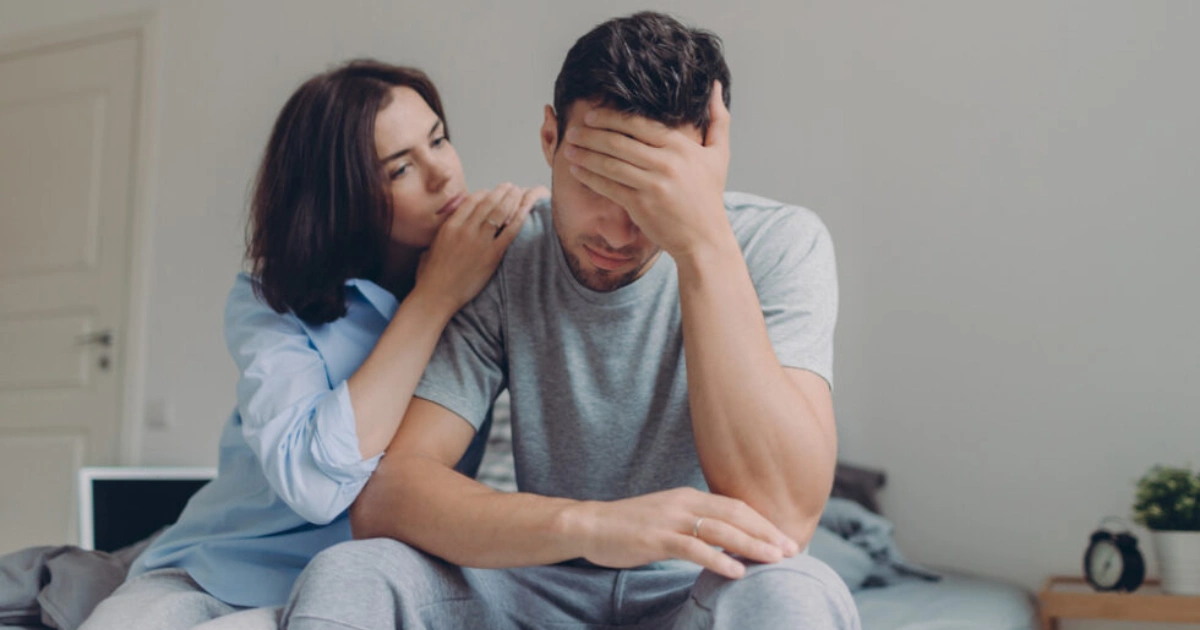 Woman comforting a distressed man who sits with his head in his hand.