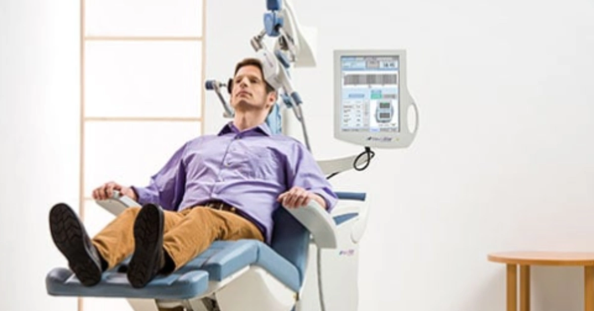 A man sits in a modern medical chair with his legs slightly raised. He appears relaxed. Medical equipment and a monitor with various controls are positioned beside him in a clean, bright room.