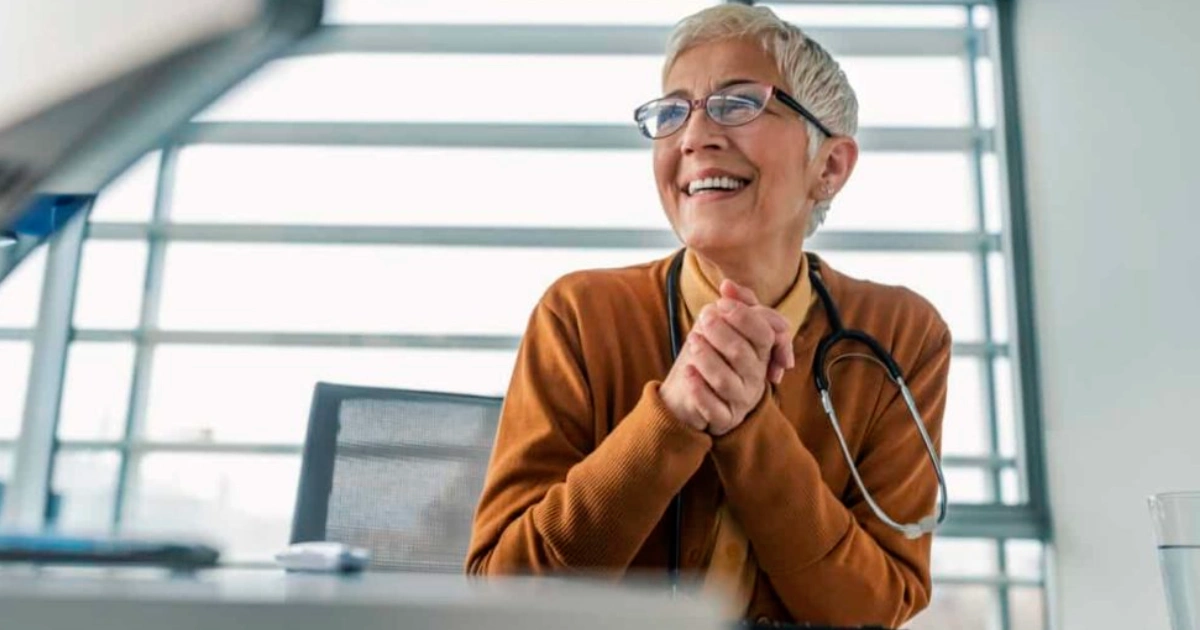 An older woman with short gray hair and glasses, wearing a brown cardigan and a stethoscope around her neck, smiles while sitting at a desk in a bright office.