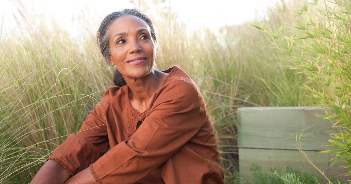 A woman with gray-streaked hair, wearing a brown top, sits outdoors among tall grasses, looking to the side and smiling softly in natural light.