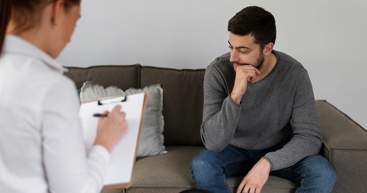 A man sits on a gray couch looking thoughtful, with his hand on his chin, while a person holding a clipboard and pen listens and takes notes during a therapy or counseling session.