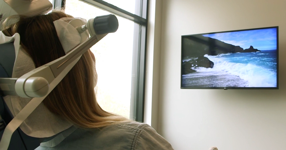 Patient receiving TMS treatment while watching a calming ocean scene on a wall-mounted screen.