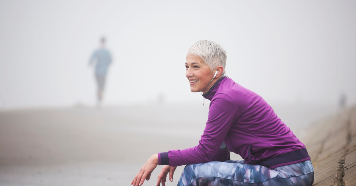 A smiling woman with short gray hair, wearing a purple jacket and patterned leggings, sits on a concrete ledge outdoors on a foggy day. She is listening to earphones, with a blurry figure jogging in the background.