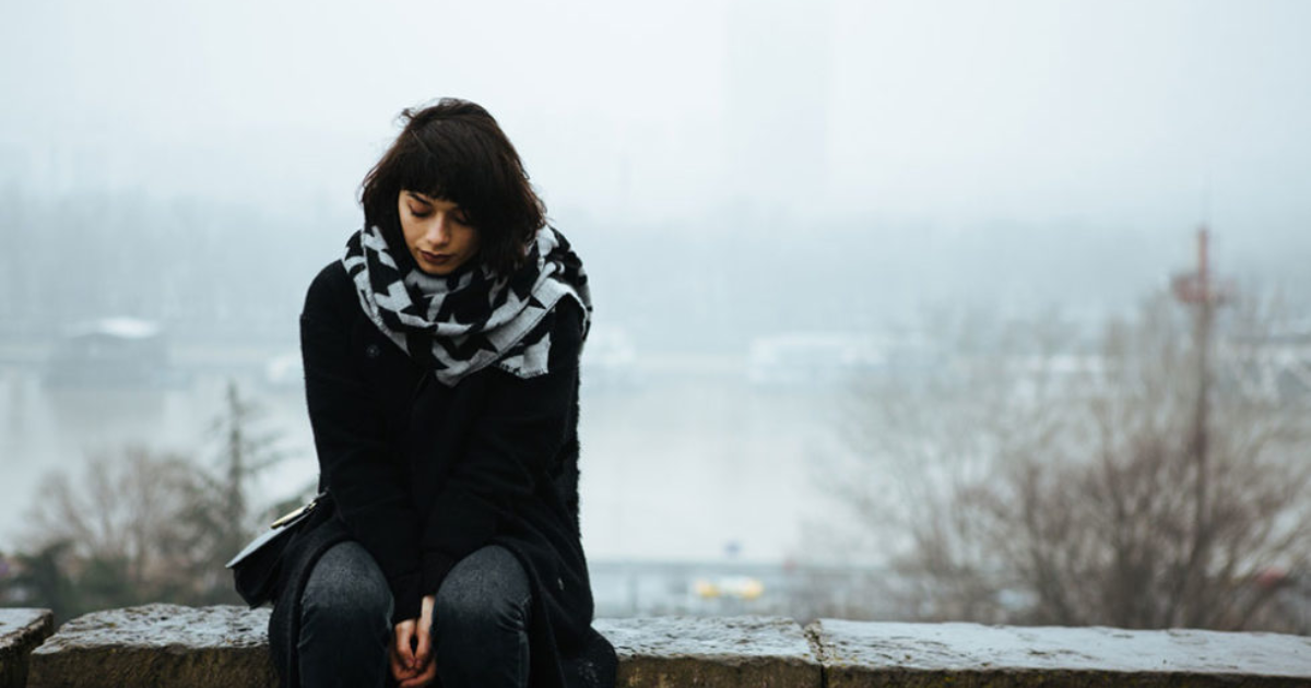 A young woman sits alone on a stone ledge outdoors, looking down with a thoughtful or sad expression. She is wearing a black coat and a patterned scarf. The background is foggy and shows blurred trees and a river.