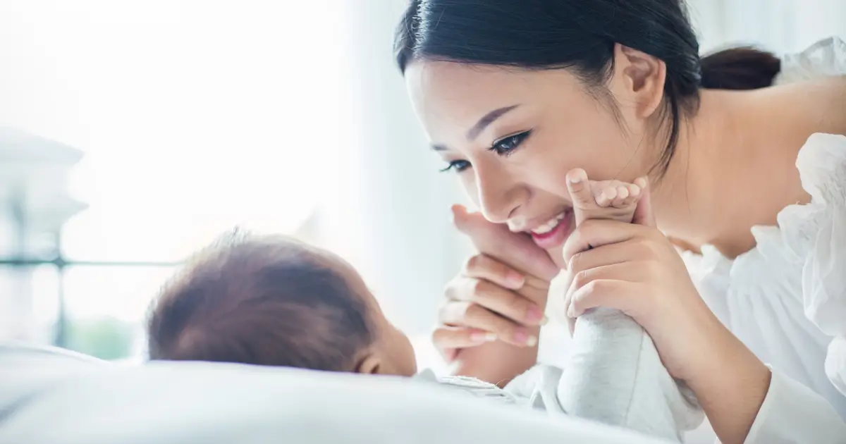 Mother smiling warmly while holding her baby’s hands during a close, affectionate moment.